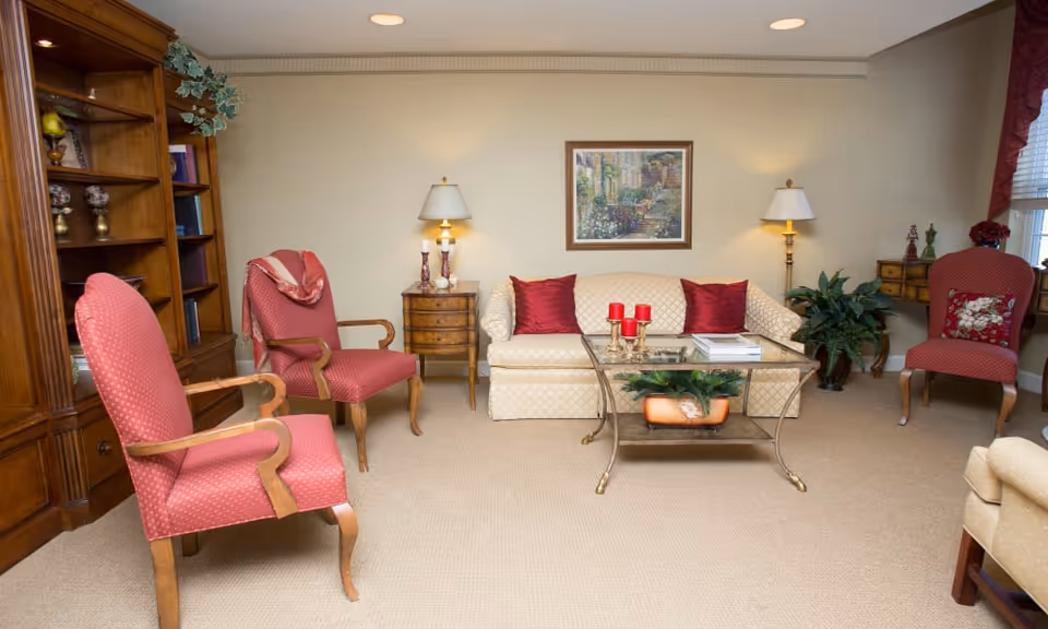 A cozy living room with a beige patterned sofa adorned with red pillows, two red upholstered armchairs with wooden arms, a glass coffee table with red candles and a plant underneath, two side tables with lamps, a wooden bookshelf, a framed painting on the wall, and a window with red curtains.