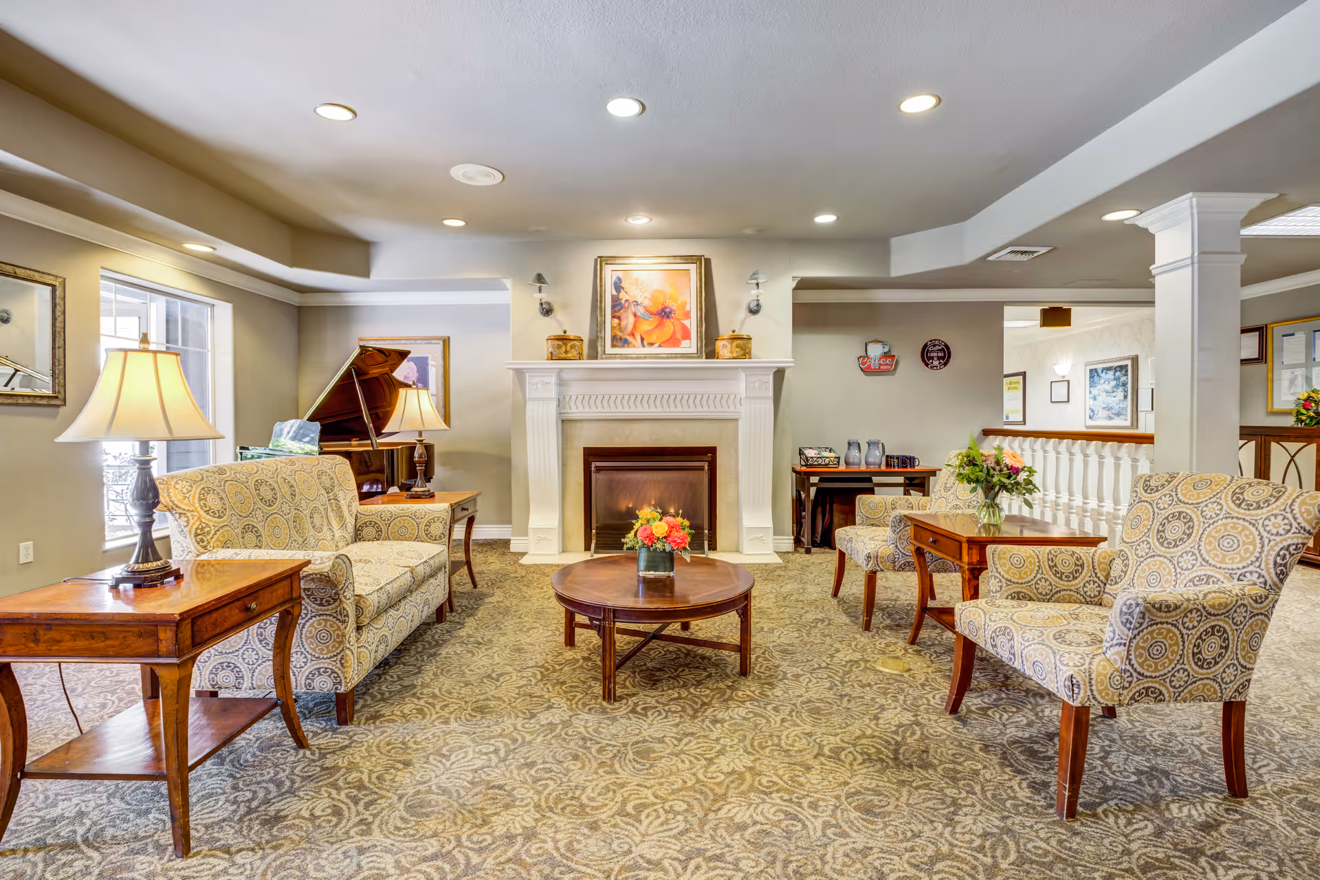 A cozy living room area in a senior living facility featuring patterned armchairs and a sofa arranged around a wooden coffee table with a flower arrangement. There is a white fireplace with a floral painting above it, two table lamps on side tables, and a grand piano in the corner. The room has recessed ceiling lights and a patterned carpet.