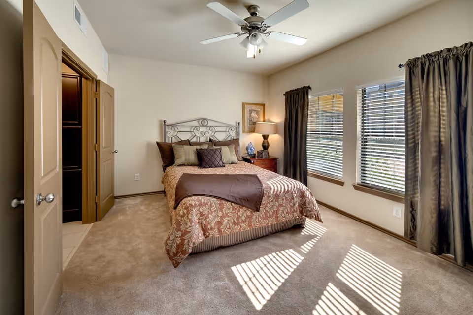 Sunlit bedroom with a decorative bed, nightstand, ceiling fan, and three windows with blinds and curtains.