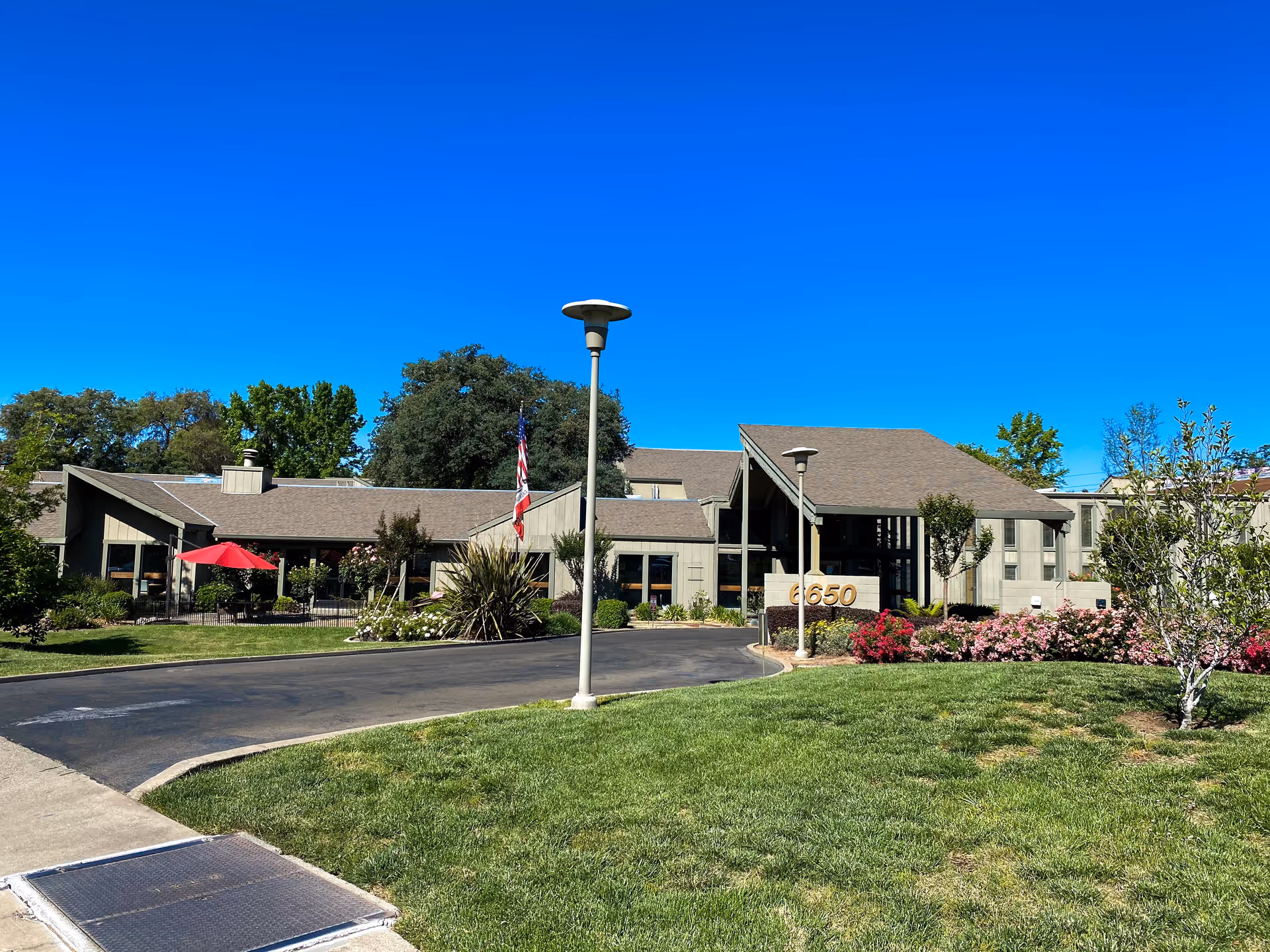 Exterior view of The Crest at Citrus Heights facility showing a single-story building with a sloped roof, surrounded by green lawns, trees, and colorful flower beds under a clear blue sky. There is a driveway leading to the entrance, a flagpole with an American flag, and a sign displaying the number 6650.