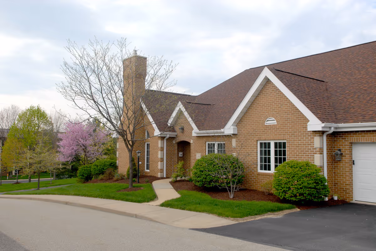 Exterior view of a brick building with a brown shingled roof, a chimney, and white-trimmed windows. There is a curved sidewalk leading to the entrance, surrounded by green grass, bushes, and trees, some with pink blossoms. The sky is partly cloudy.