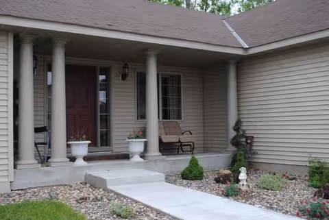 Front entrance of a senior living facility with columns, chairs, and landscaped walkway.