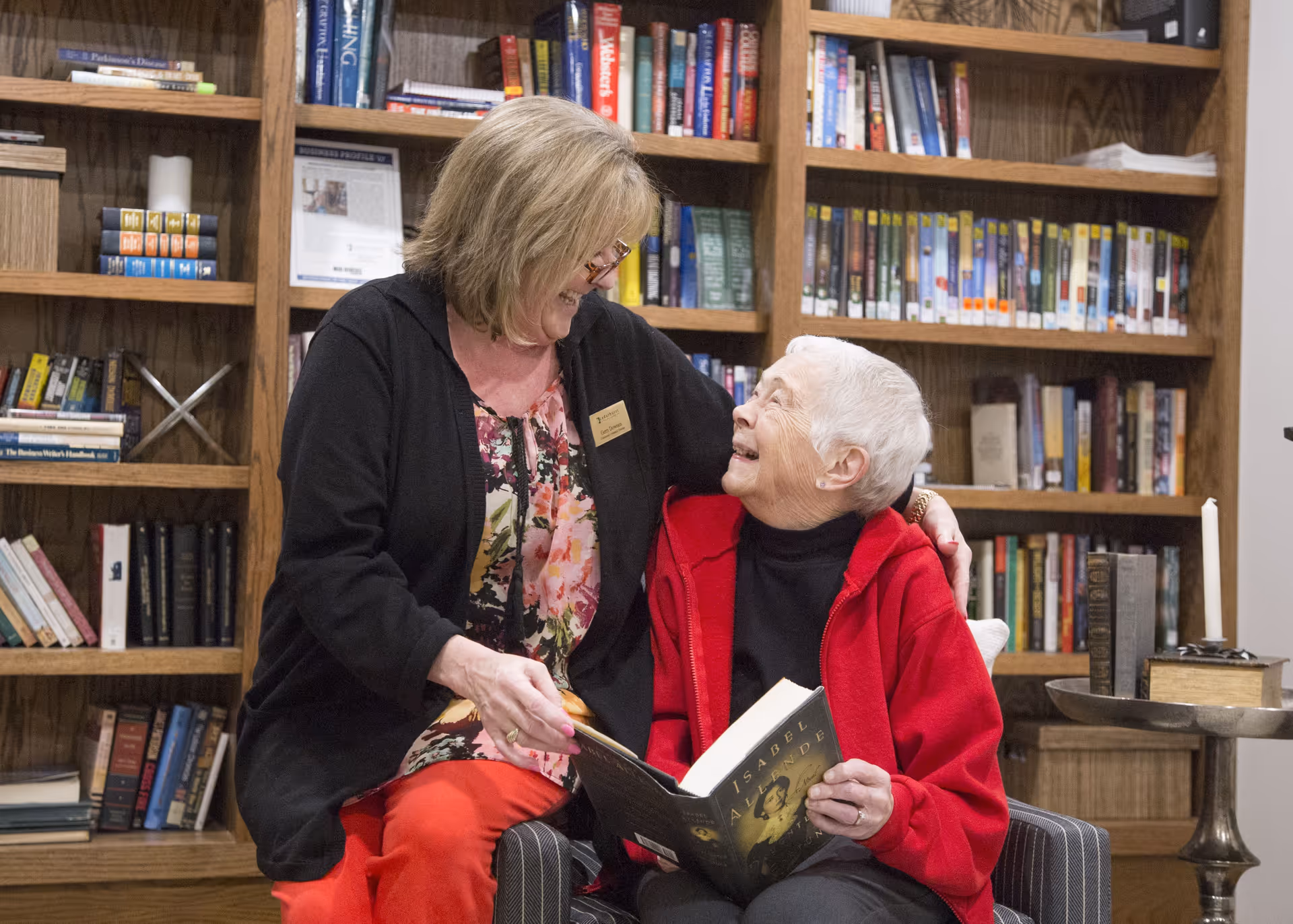 An elderly woman in a red jacket sitting in a chair holding a book, smiling and looking up at a woman standing next to her who is also smiling and wearing glasses, a floral top, and a black cardigan. They are in a room with wooden bookshelves filled with books in the background.