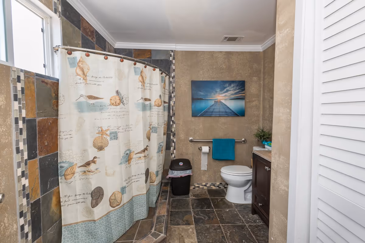 A bathroom with a tiled floor and walls, featuring a shower with a curtain decorated with seashells and birds. There is a white toilet with a teal towel hanging on a grab bar above the toilet paper holder. A small black trash bin is next to the toilet. A framed picture of a pier extending into a calm body of water at sunset hangs on the wall above the towel. A wooden vanity with a countertop and a small plant is visible on the right side.