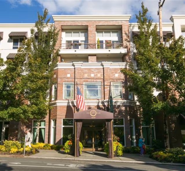 Front exterior view of a multi-story brick building with a covered entrance canopy displaying the name University House Wallingford. The building is flanked by tall trees and has American and other flags near the entrance. There are some plants and flowers around the entrance area and a person walking on the sidewalk.