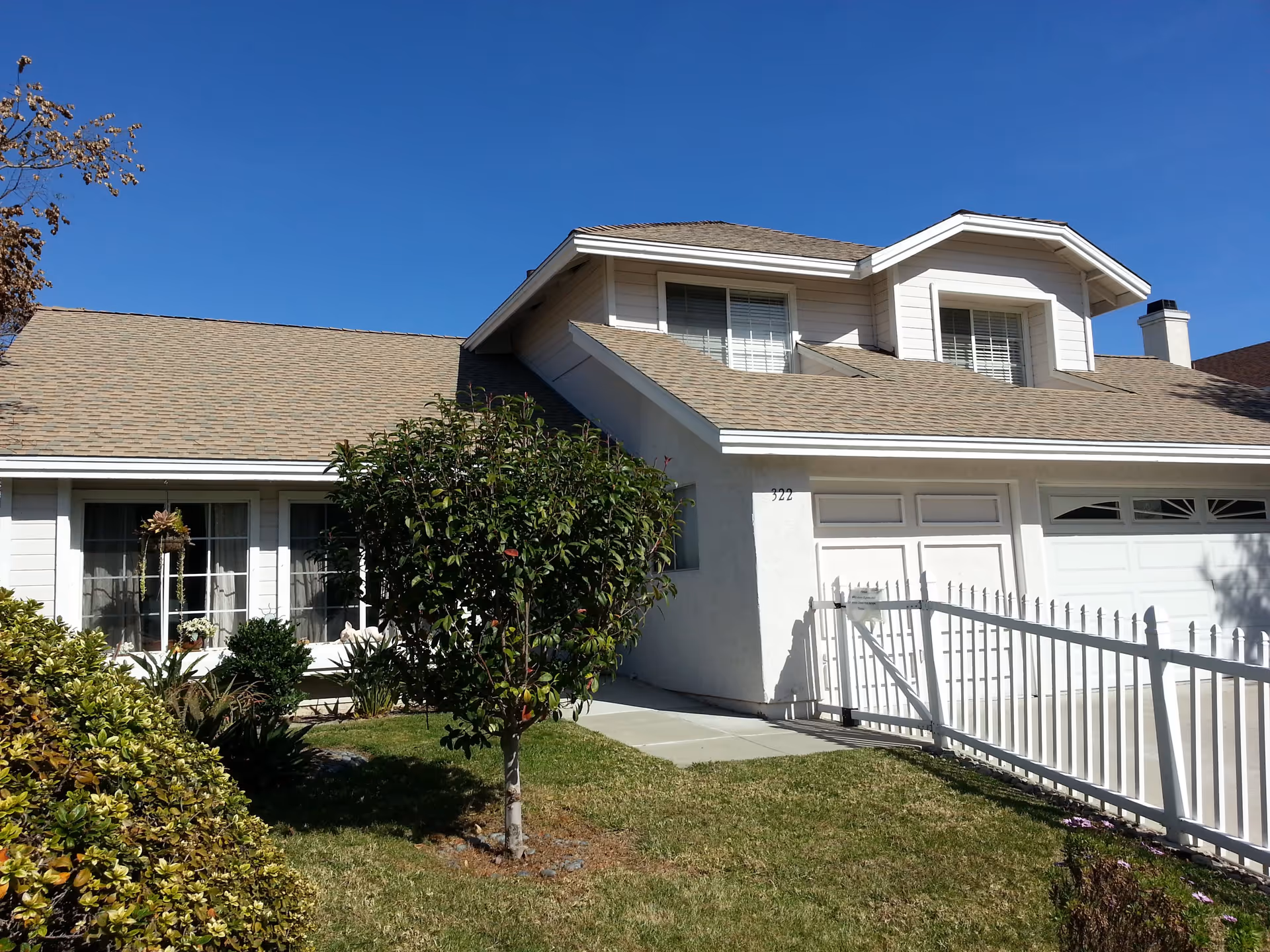 Exterior view of a two-story residential building with beige siding and a brown shingle roof under a clear blue sky. The front yard has green grass, bushes, and a small tree. There is a white fence and a driveway leading to a garage with two white doors.