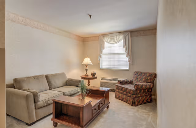 Small living room featuring a beige sofa, patterned armchair, wooden coffee table, side table with a lamp, and a window with blinds and drapery.