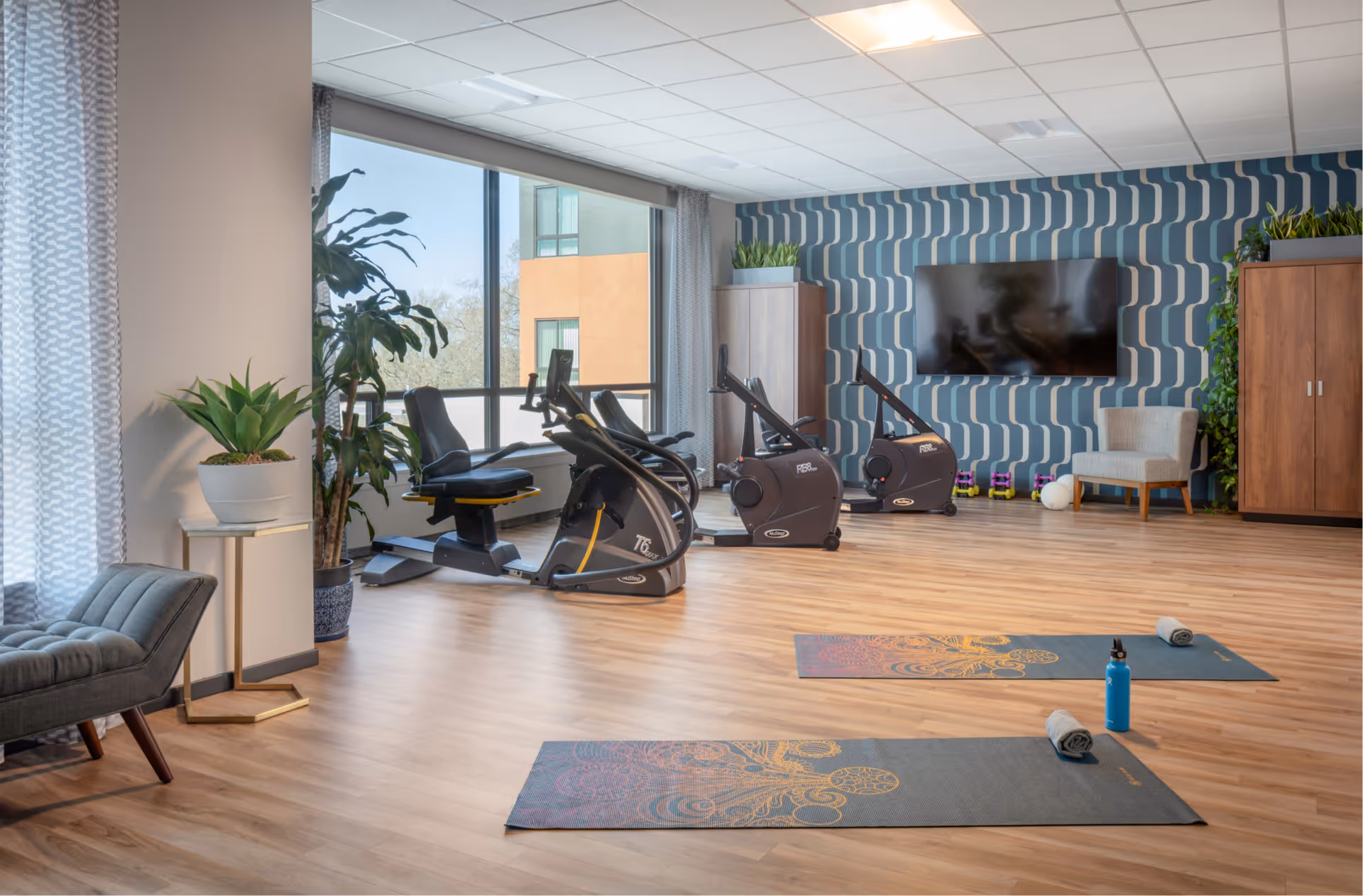 A bright and spacious fitness room with large windows letting in natural light. The room features two exercise bikes, two yoga mats with towels and a water bottle, a comfortable chair, potted plants, wooden cabinets, and a large flat-screen TV mounted on a patterned blue and white wall.