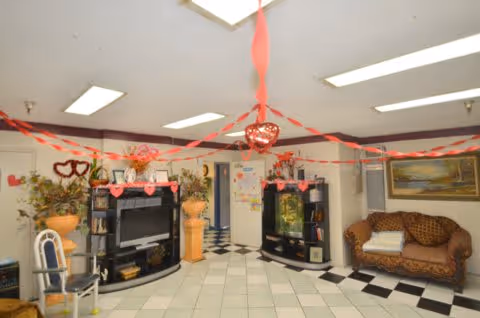 A decorated common area with white tiled floor and black accents, featuring two black entertainment centers with a TV and fish tank, a brown patterned sofa with folded towels, and chairs. Red and pink heart-shaped decorations and streamers hang from the ceiling, suggesting a Valentine's Day theme.
