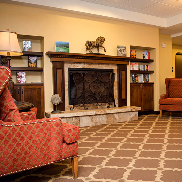 A cozy senior living room with a decorative fireplace featuring a wrought iron screen. The room has patterned carpet flooring, two red upholstered armchairs with a diamond pattern, built-in wooden shelves with books and decorative items, and a table lamp on the left side.