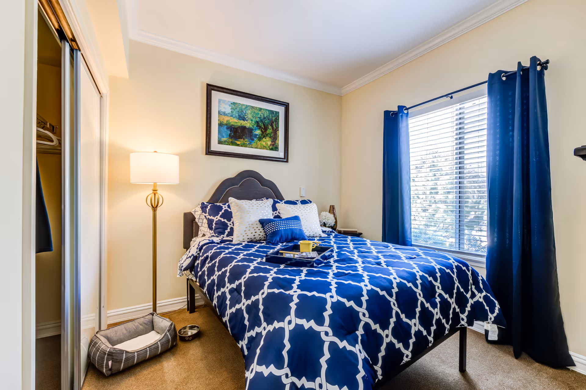 A cozy bedroom with a bed dressed in blue and white patterned bedding, several pillows, and a tray with a yellow cup on it. There is a floor lamp next to the bed, a framed painting on the wall above the headboard, a window with blue curtains, and a small pet bed and water bowl on the floor near a mirrored closet door.
