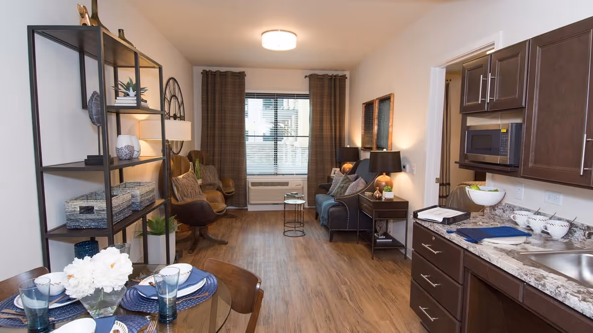 Interior view of a senior living apartment featuring a dining area with a glass table set with plates, bowls, and glasses, a living area with a sofa, chairs, side tables with lamps, and a window with brown curtains. The kitchen area has dark wood cabinets, a microwave, a sink, and a countertop with a bowl of fruit and cups.