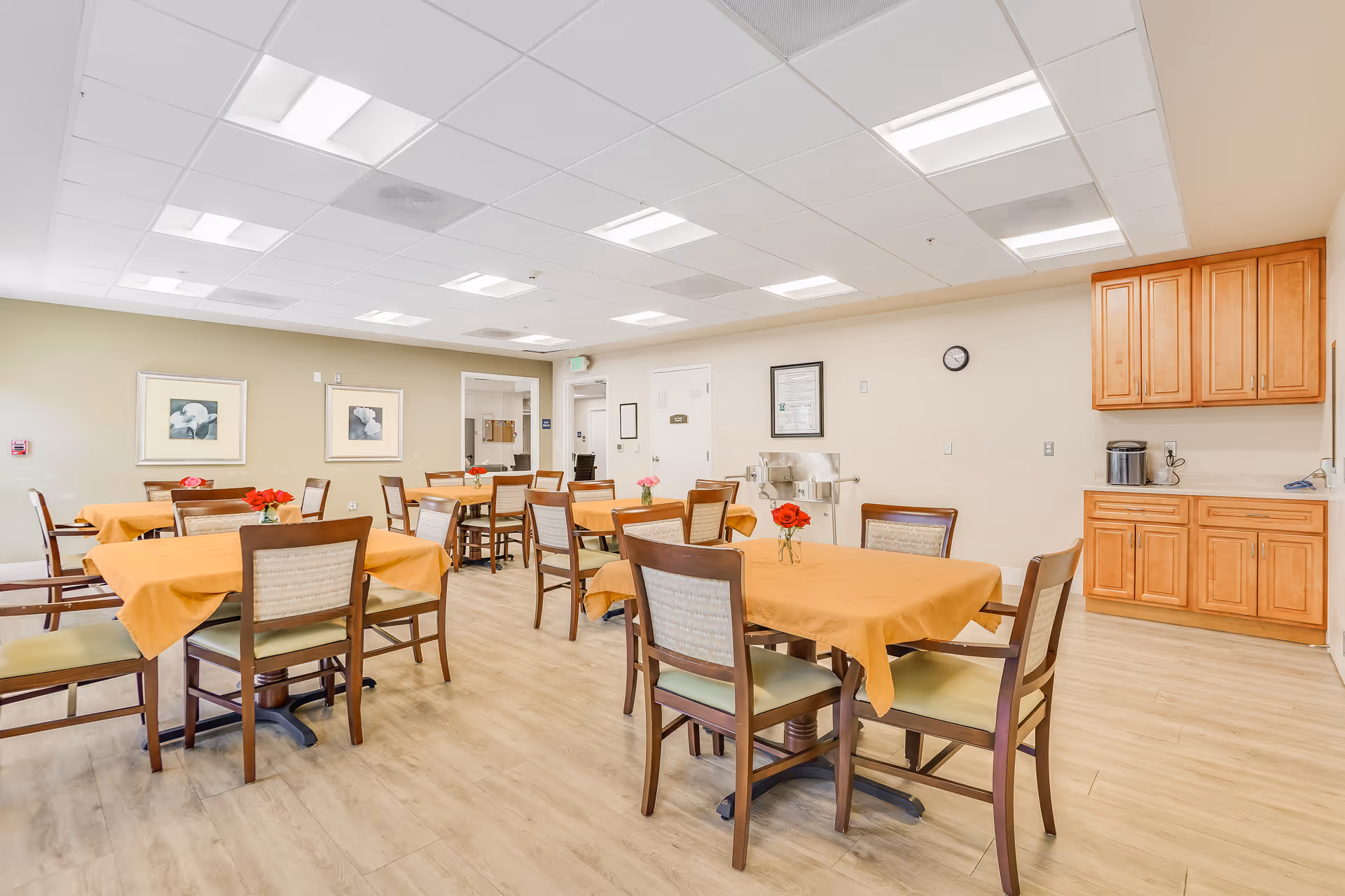 A bright dining room with several wooden tables covered with orange tablecloths, each table surrounded by wooden chairs with cushioned seats. Small vases with red flowers are placed on the tables. The room has light-colored walls, framed artwork, a clock, and wooden cabinets along one wall. The floor is light wood, and the ceiling has recessed lighting panels.