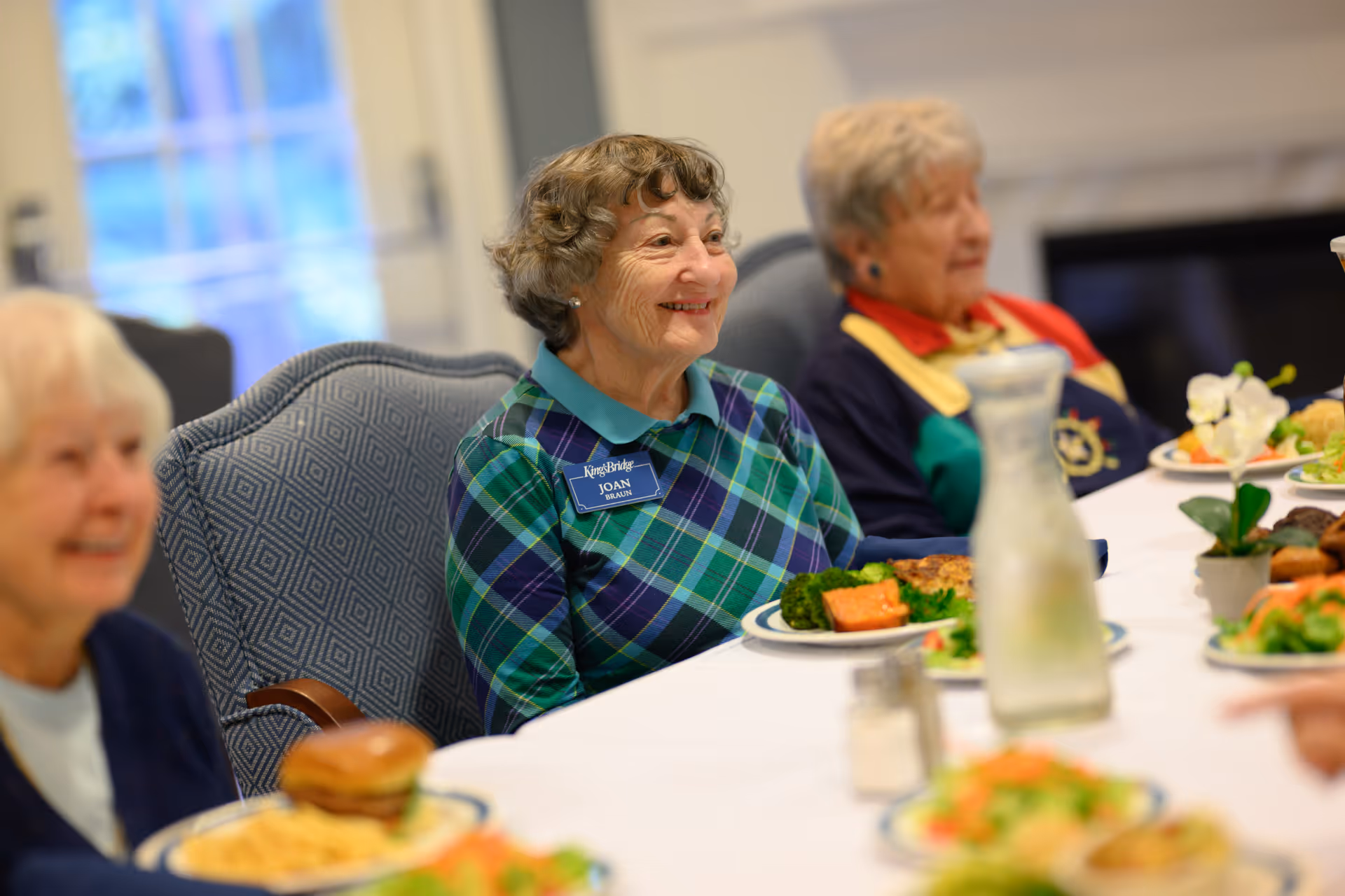 Elderly residents smiling while seated at a communal dining table with plates of food.