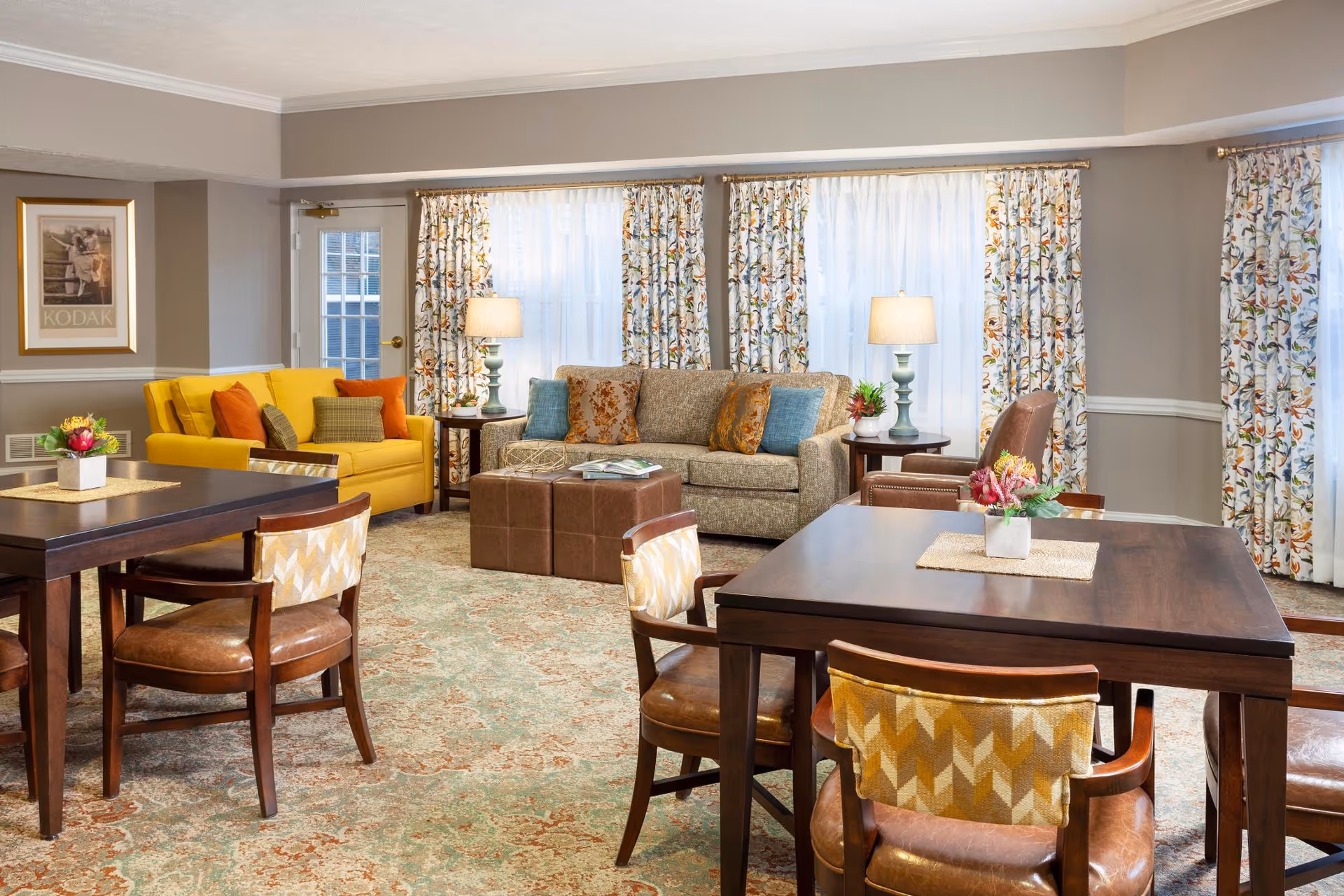 A cozy living room area with two dark wooden tables surrounded by chairs with patterned cushions. In the background, there is a yellow loveseat with orange and green pillows, a beige sofa with blue and orange pillows, two side tables with lamps, and floral patterned curtains covering large windows. The room has a patterned carpet and neutral-colored walls with a framed Kodak poster on one side.
