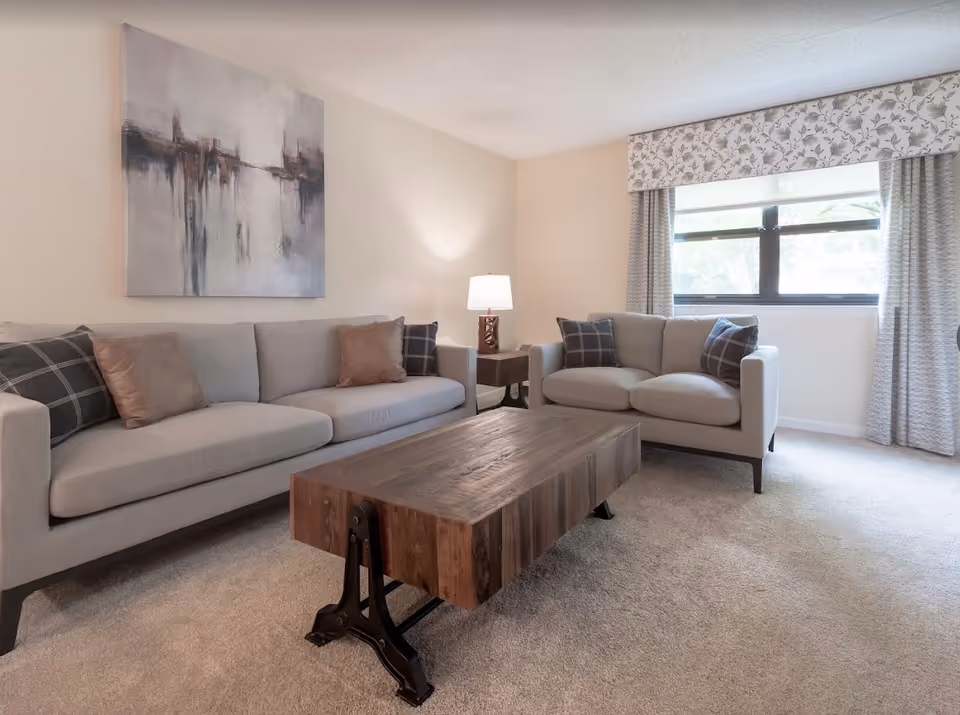 A cozy living room with two light gray sofas adorned with plaid and solid pillows, a rustic wooden coffee table with metal legs, a side table with a lamp, a large window with floral valance and curtains, and an abstract painting on the wall.