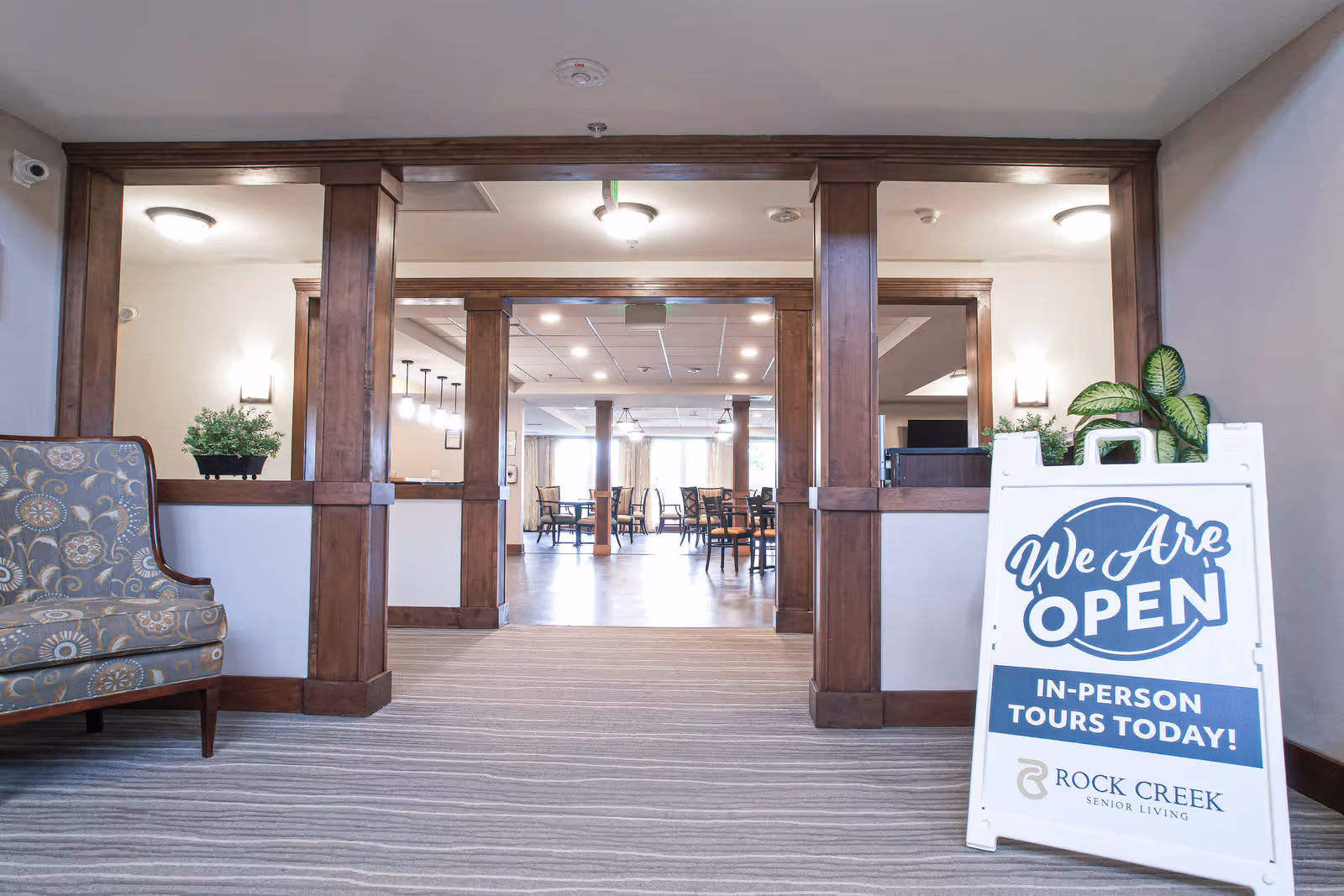 Lobby-style entrance into a senior living common area with wooden columns, visible dining tables and a white sandwich board sign reading 'We Are Open'.