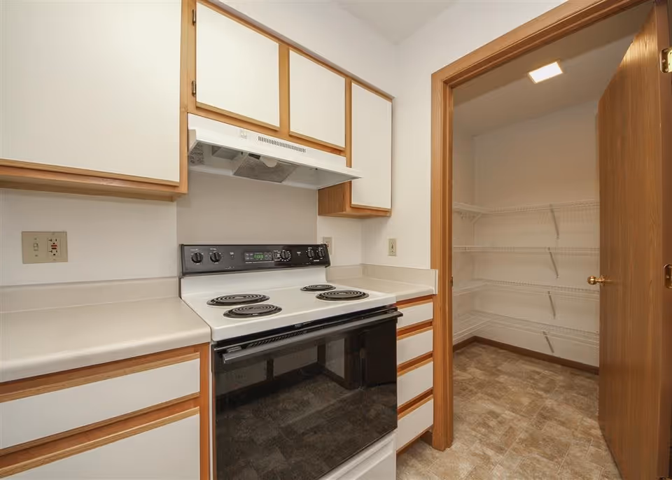 Interior view of a kitchen featuring an electric stove with four coil burners, white countertops, and wooden cabinets with white doors. To the right, there is an open door leading to a pantry with multiple wire shelves and a tiled floor.