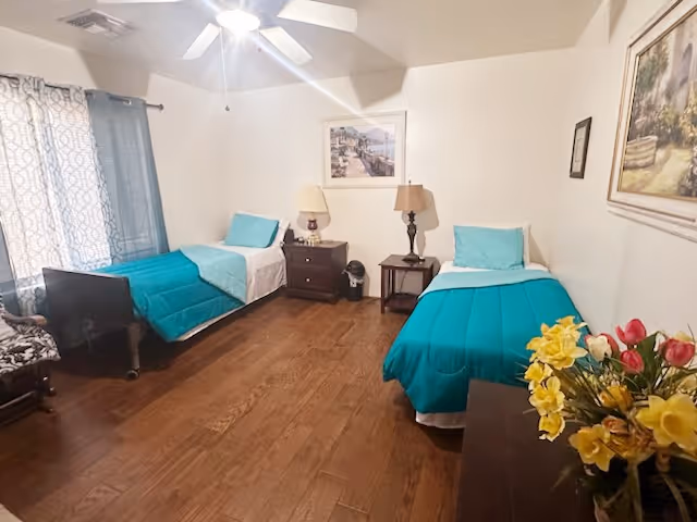 A bedroom with two single beds, each with teal bedding and pillows. Between the beds are two small nightstands with lamps. The room has wooden flooring, a ceiling fan with a light, and a window with patterned curtains. There are framed pictures on the walls and a vase with yellow and red flowers on a dresser in the foreground.