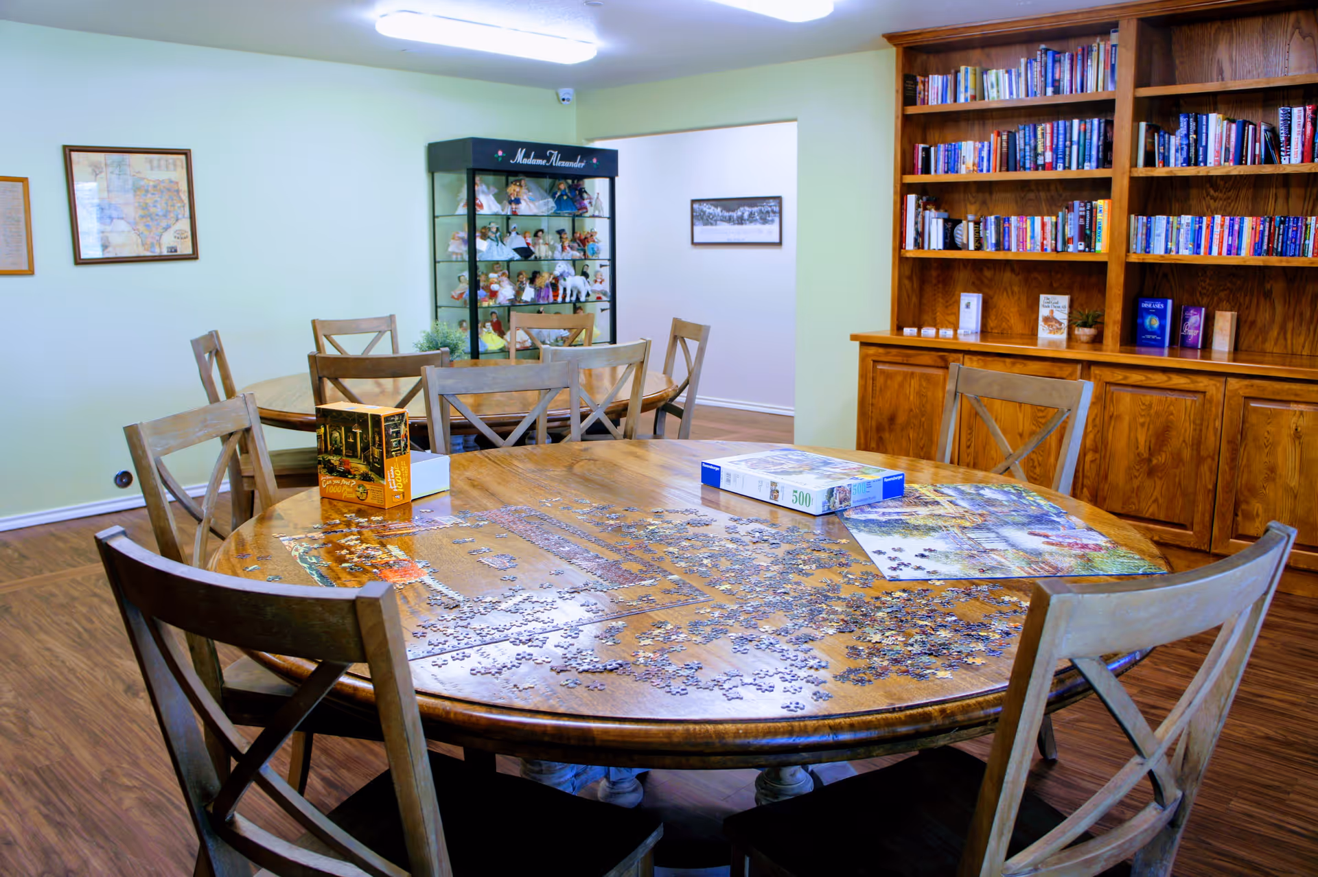 A cozy room with a large round wooden table surrounded by wooden chairs. On the table, there is an incomplete jigsaw puzzle with puzzle boxes. In the background, there is a wooden bookshelf filled with books and a glass display cabinet with dolls. The room has light green walls and wooden flooring.