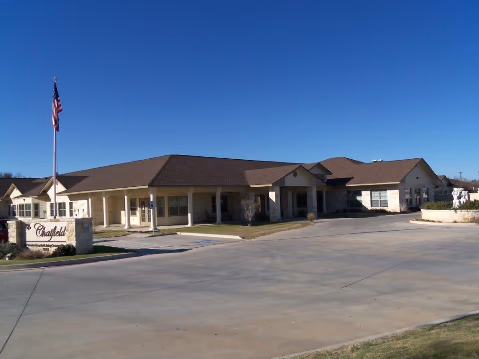 Single-story assisted living building with a covered entrance, an American flag, and a 'Chatfield' sign in front under a clear blue sky.