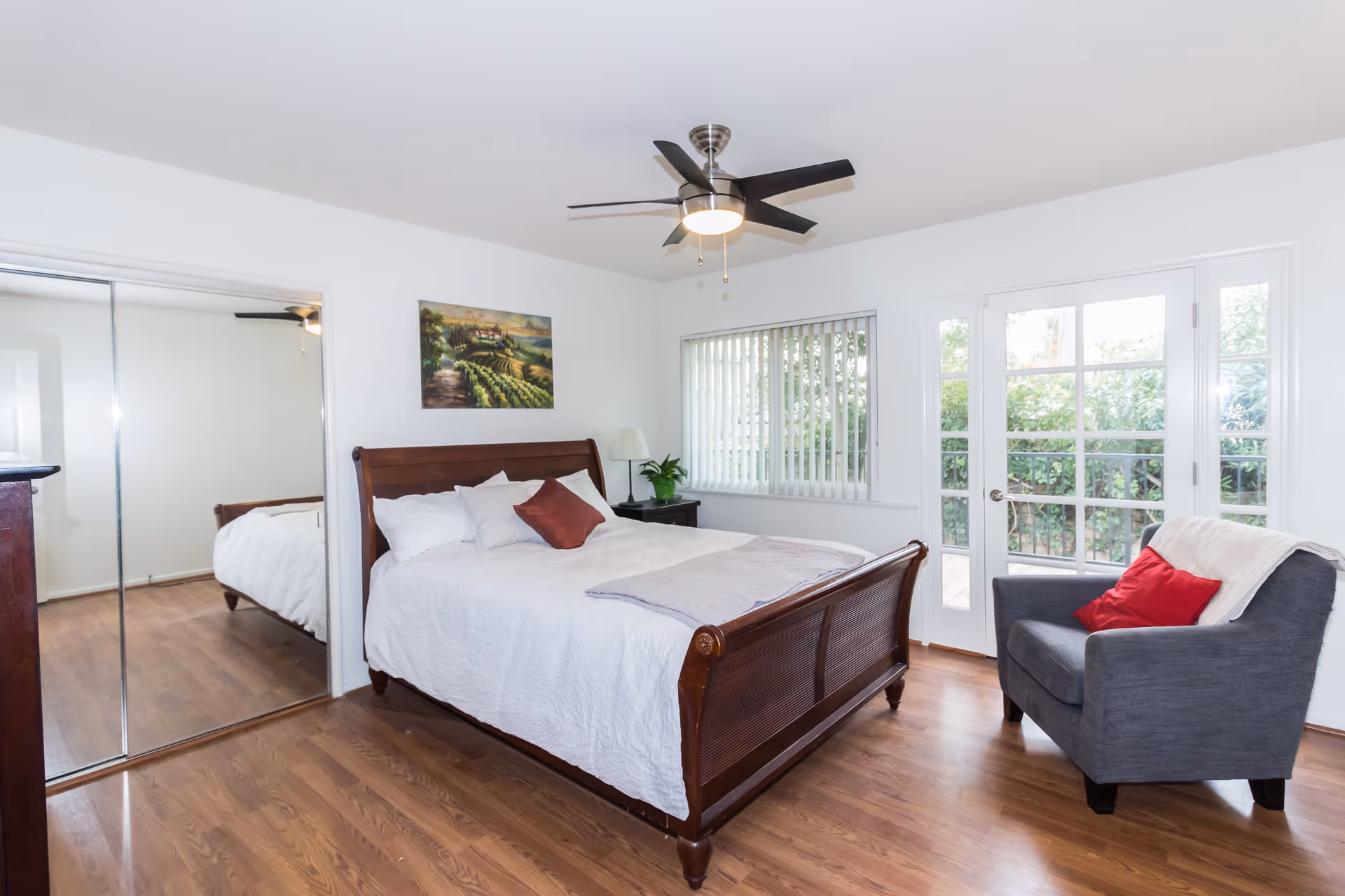 A bright bedroom with a wooden bed frame, white bedding, and several pillows. There is a gray armchair with a red pillow and a white throw blanket near glass doors leading outside. A ceiling fan with a light is mounted on the ceiling, and a painting hangs on the wall above the bed. The room has wooden flooring and a window with vertical blinds.