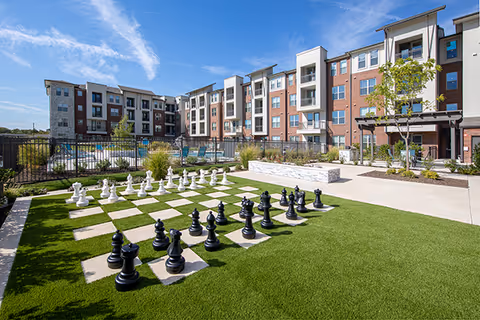 Outdoor courtyard with a giant chessboard on artificial turf, a pool area, and a multi-story apartment building under a blue sky.