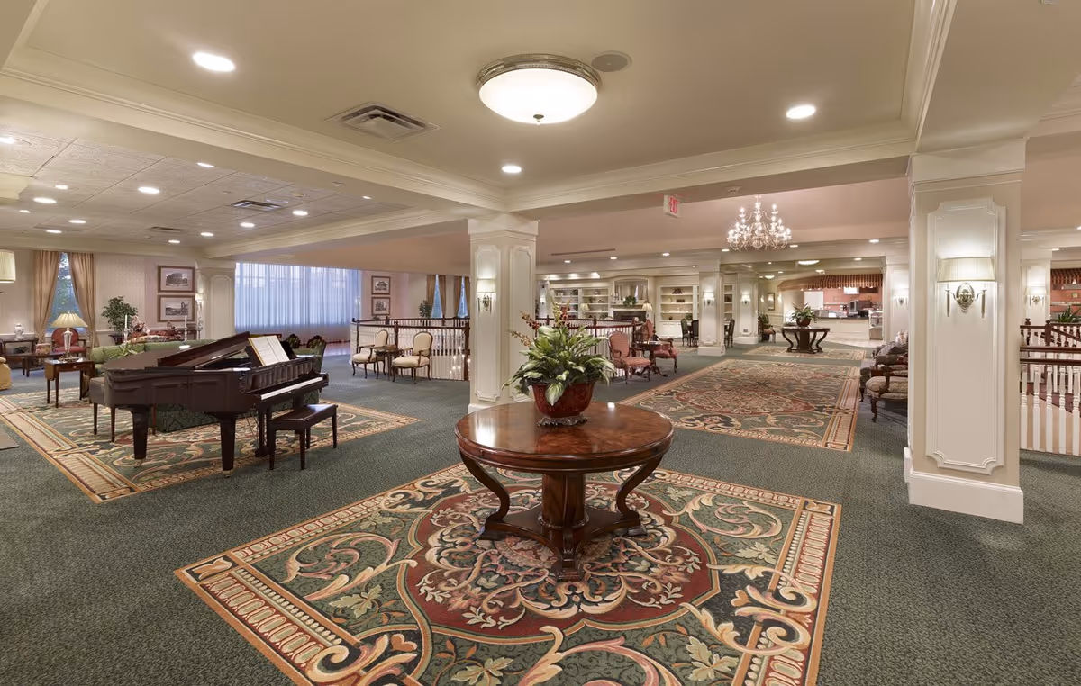 Spacious senior living facility common area with a grand piano, seating areas, decorative rugs and a central table with a plant.