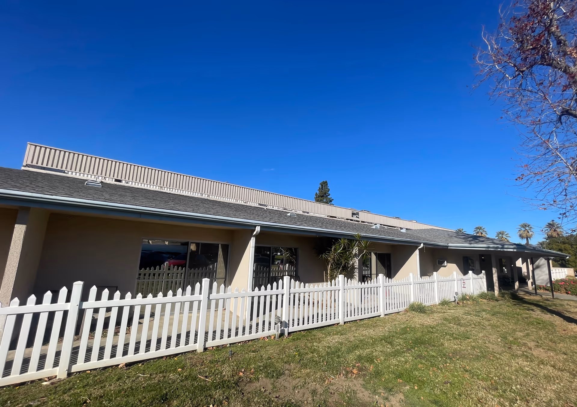 Single-story beige building with a white picket fence and grassy lawn under a clear blue sky.