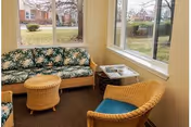 A cozy sitting area with wicker furniture including a floral-patterned sofa, a wicker chair with a blue cushion, and a small round wicker table. There is a side table with magazines next to the sofa. Large windows provide a view of the outdoor garden and neighboring buildings.