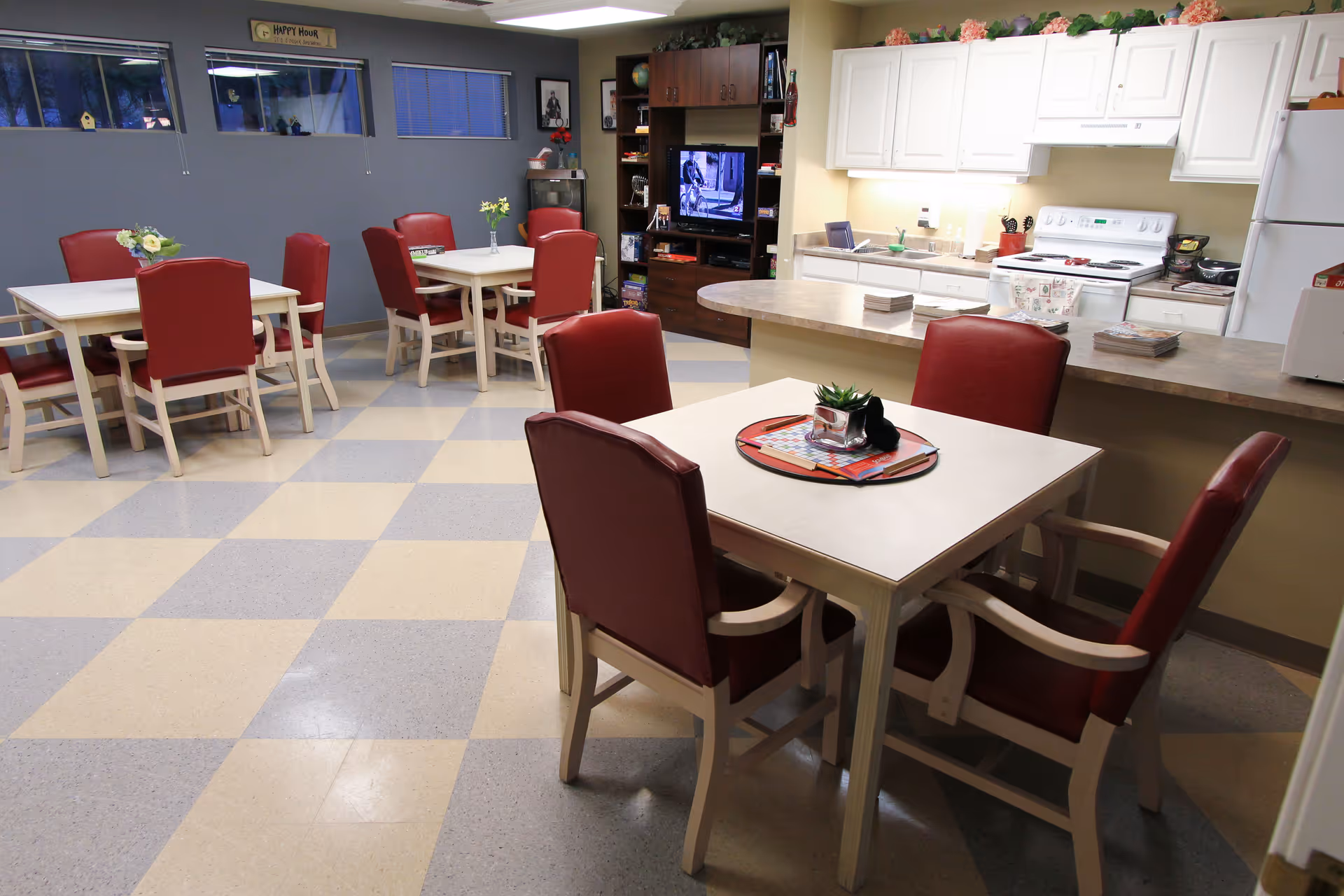 A communal dining and kitchen area with several square tables surrounded by red cushioned chairs. The kitchen has white cabinets, a stove, and a countertop with some items on it. In the background, there is a TV on a wooden stand and some shelves with books and decorations. The floor has a checkered pattern in beige and gray.
