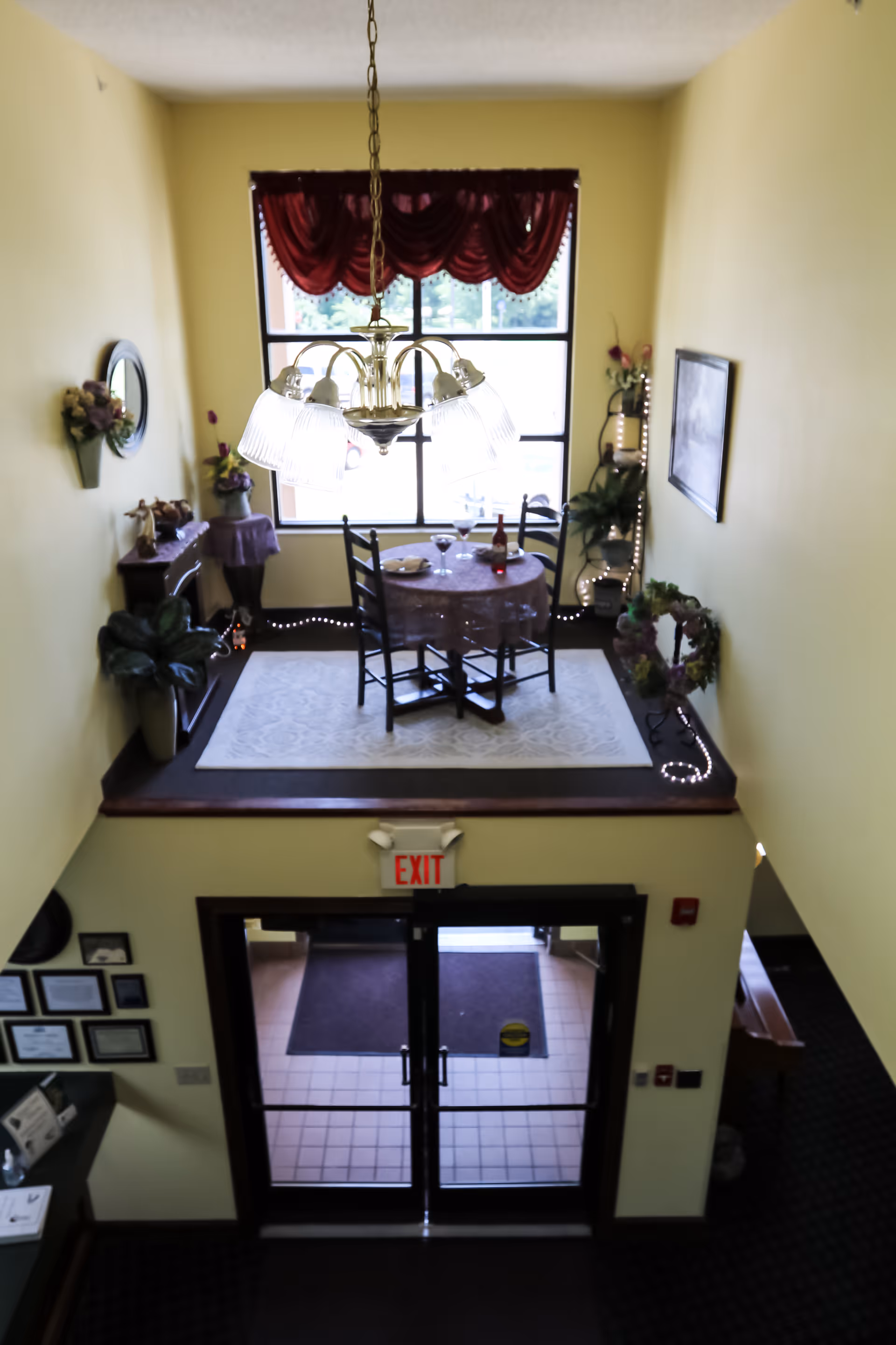 View from above of a small dining area with a round table covered with a tablecloth, set with plates, glasses, and a bottle of wine. The dining area is decorated with plants, flowers, a mirror, and a framed picture, illuminated by a chandelier. Below the dining area is a double glass door exit with an exit sign above it.