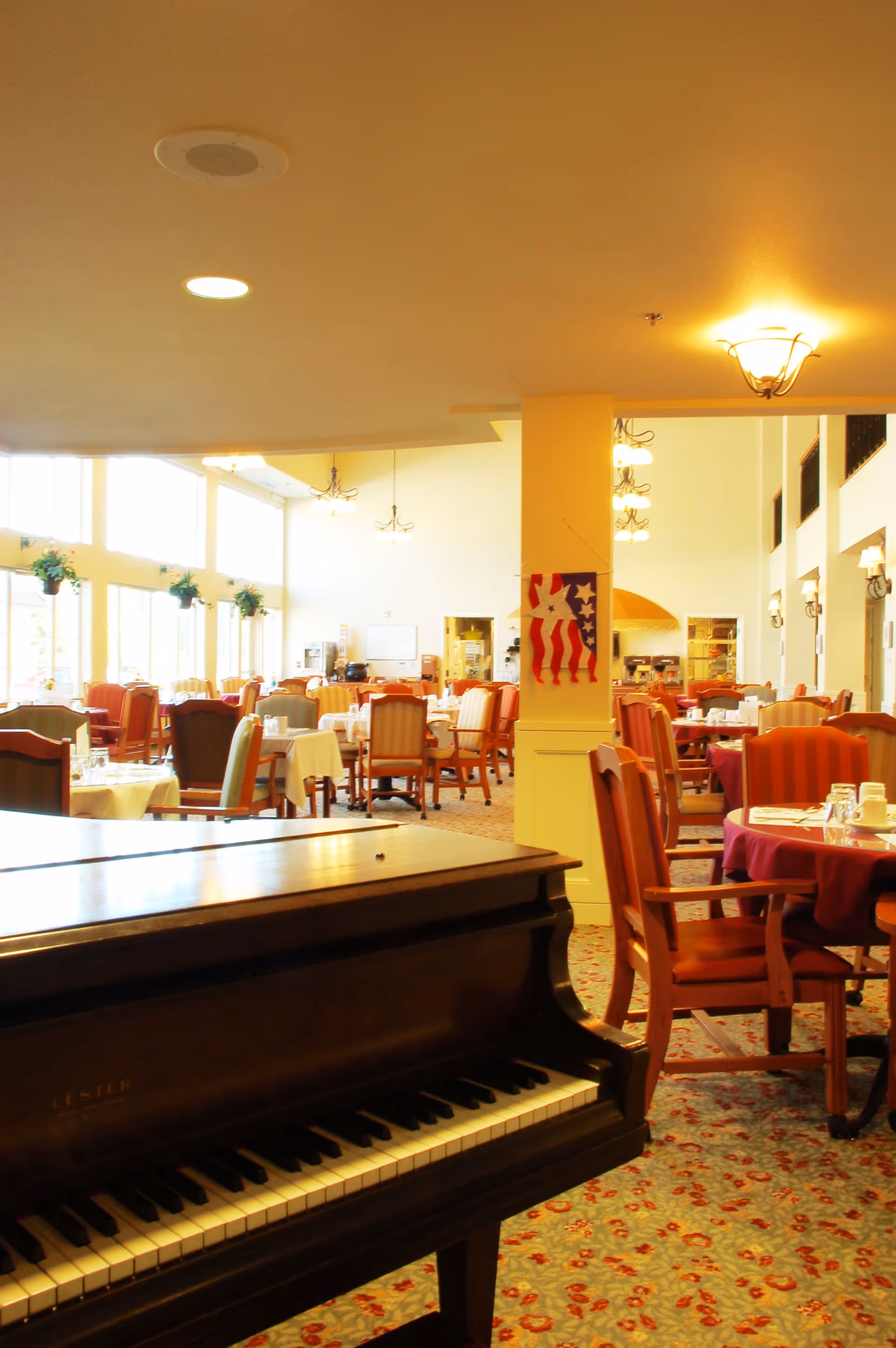 Large dining room with a grand piano in the foreground, multiple set tables and chairs, tall windows, and soft overhead lighting.