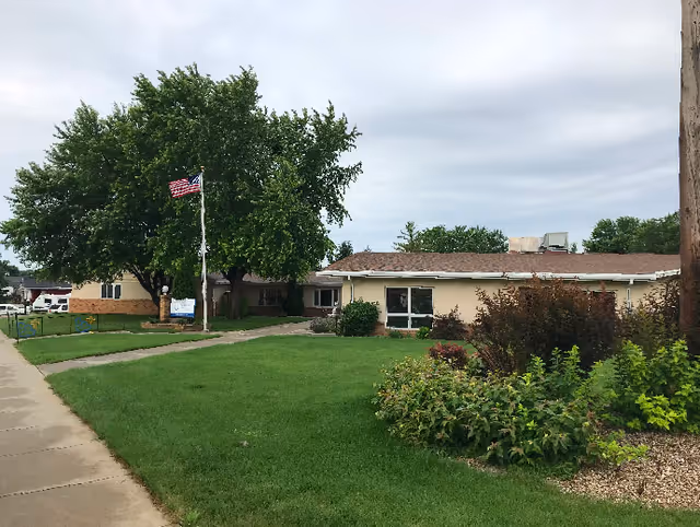 Exterior view of a single-story building with a brown roof and beige walls, surrounded by green grass, bushes, and trees. An American flag is flying on a flagpole near the sidewalk leading to the entrance. The sky is overcast.