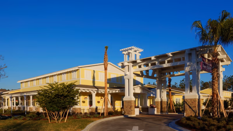 Exterior view of a senior living facility building with yellow siding and white trim, featuring a large white pergola entrance supported by stone pillars, surrounded by palm trees and landscaping under a clear blue sky.