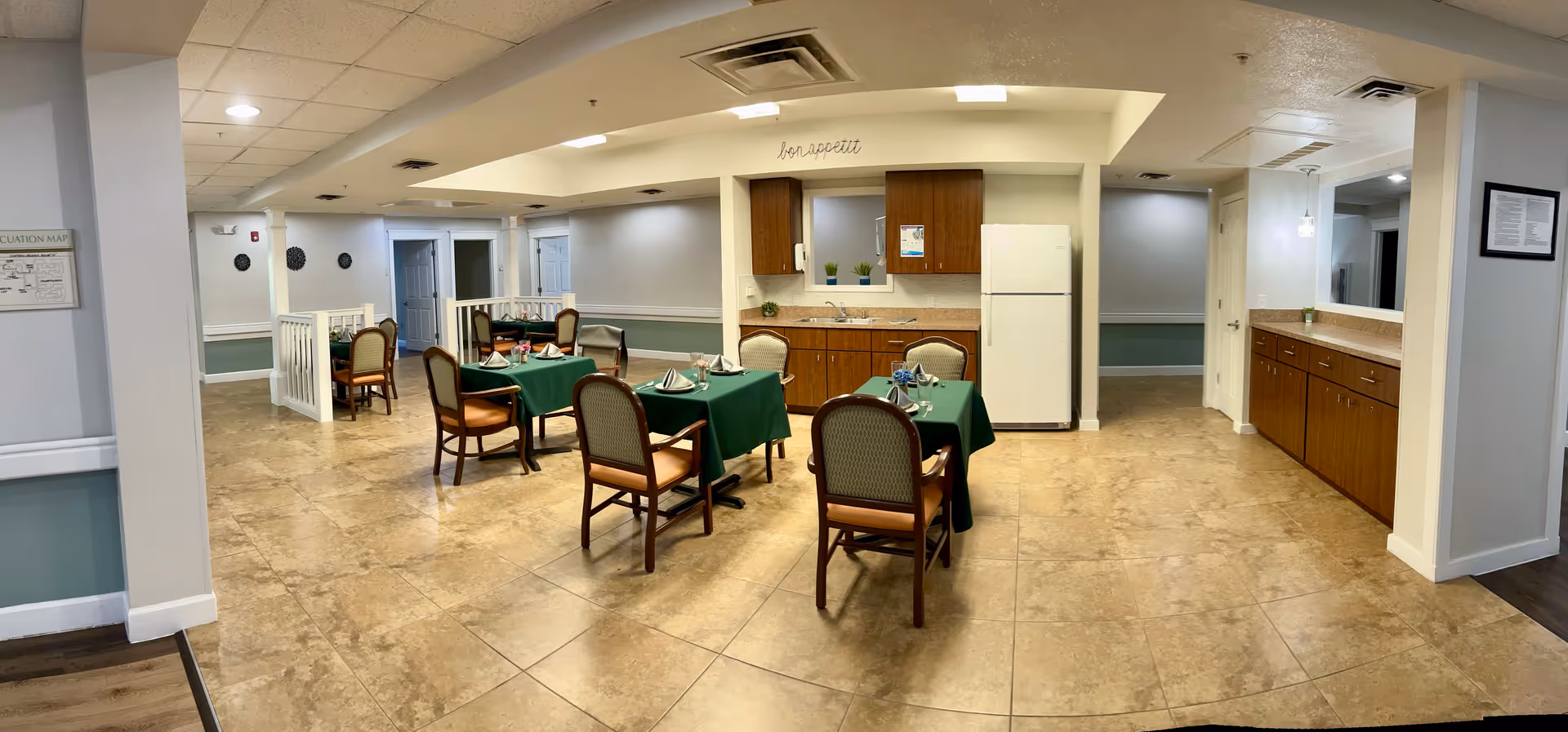 Interior view of a senior living facility dining area with several tables covered in green tablecloths, each set with napkins and glasses. The room features a small kitchenette with wooden cabinets, a white refrigerator, and a sink. The walls are painted light gray with white trim, and the floor is tiled. There are chairs around the tables and some decorative plants on the countertop and window sill.