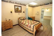 A single bed with a colorful patchwork quilt in a small, neatly arranged room. The room has light-colored walls with a decorative border near the ceiling, a wooden dresser with drawers, a framed painting above the bed, and two wall-mounted lamps. The floor is wood or laminate, and there is an open doorway leading to another room with a countertop and sink visible.