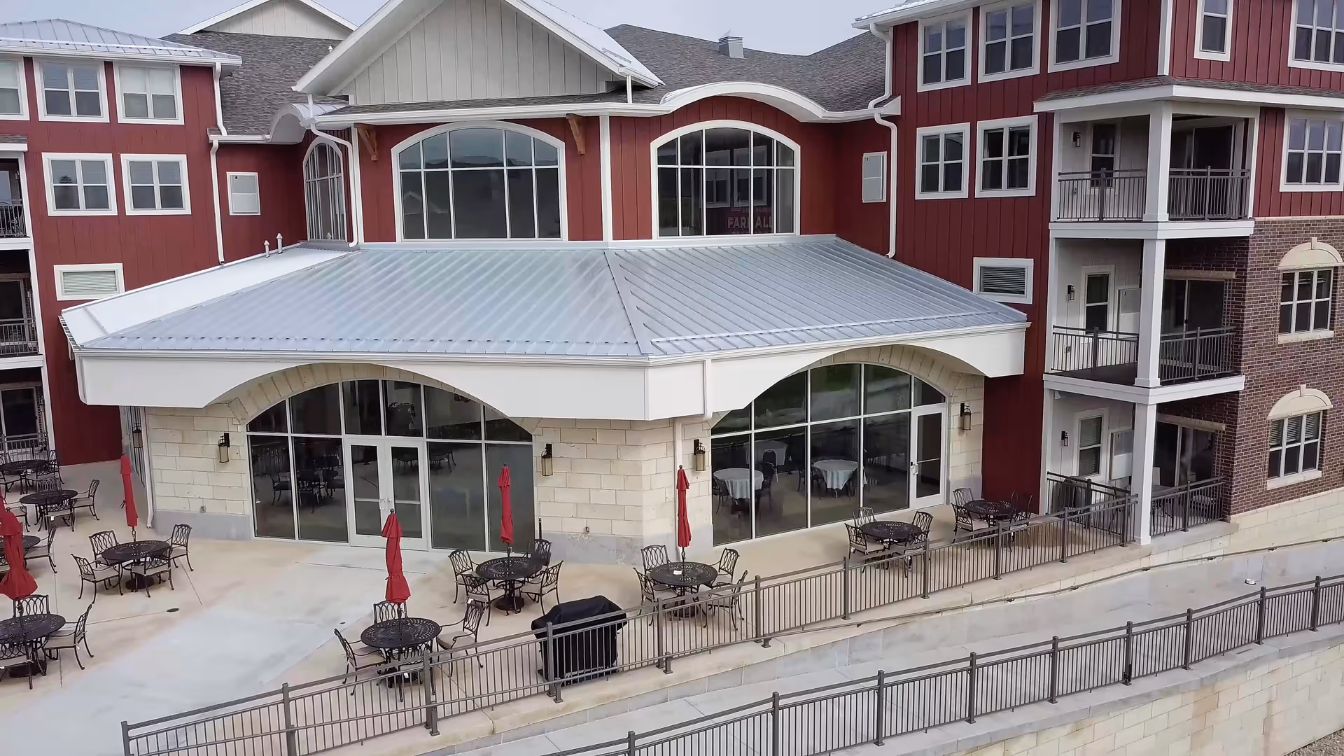 Outdoor patio area of a senior living facility with multiple round metal tables and chairs, some with closed red umbrellas. The building has large arched windows and a metal roof, with a multi-story red and beige exterior featuring balconies.