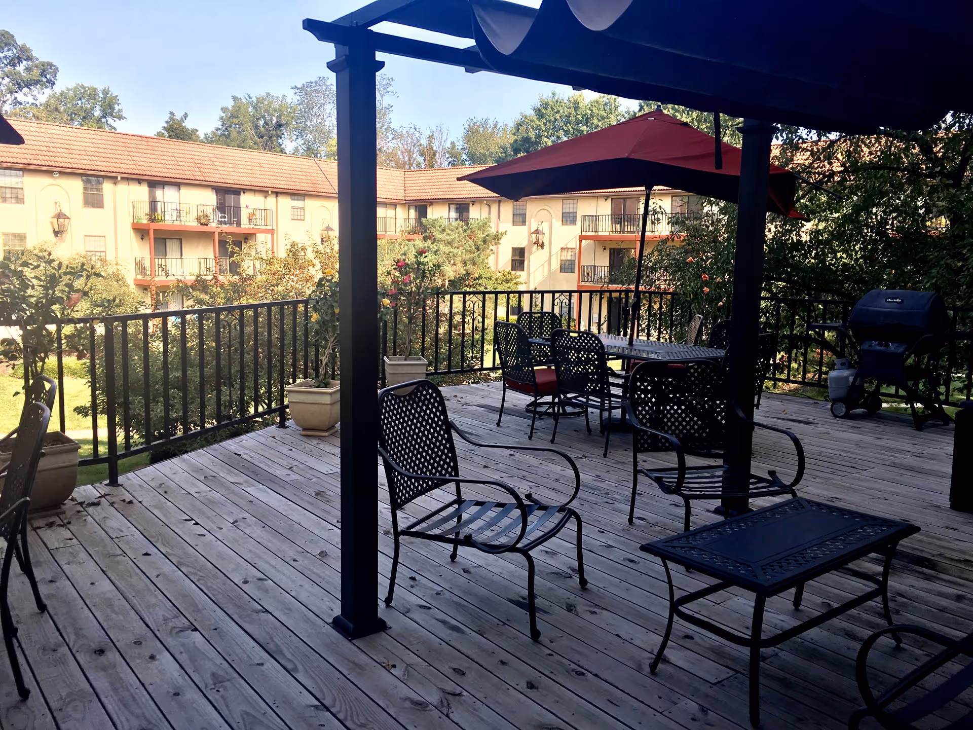 Wooden deck patio with metal chairs, tables, umbrellas and a grill overlooking a landscaped courtyard and a multi-story residential building.
