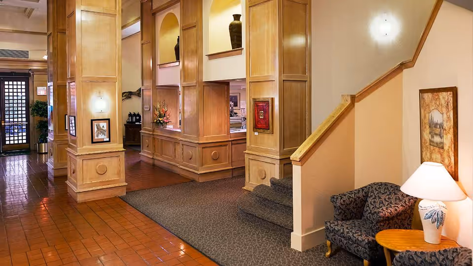 Interior view of a senior living facility lobby area with wooden paneling, tiled floor, a carpeted staircase, a patterned armchair next to a small wooden table with a lamp, framed artwork on the walls, and a reception desk in the background.