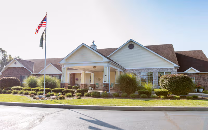 Front exterior view of a single-story senior living facility building with a covered entrance, well-maintained landscaping including bushes and grass, and an American flag on a flagpole in front.