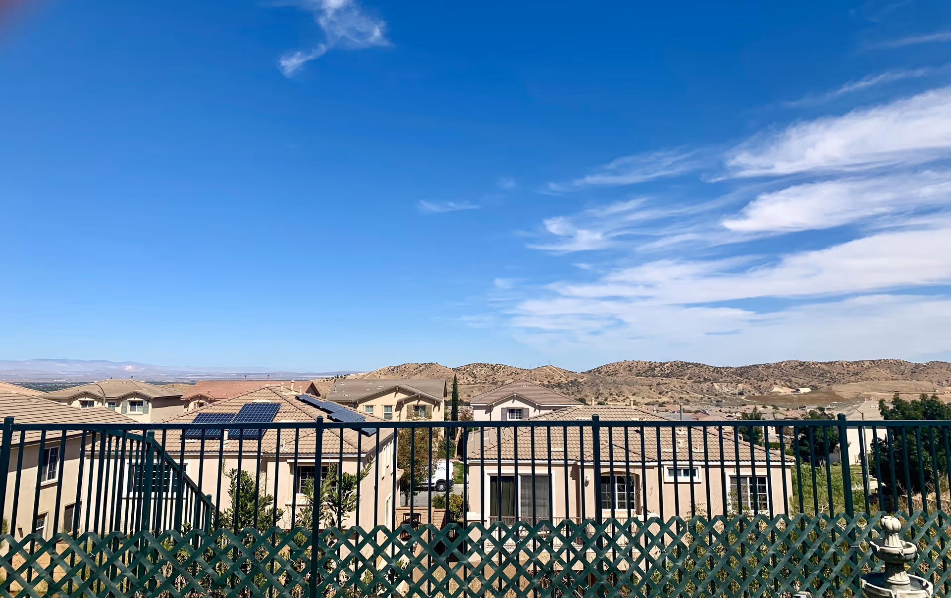 View from a balcony over tiled rooftops toward distant hills under a bright blue sky, with a railing and lattice in the foreground.