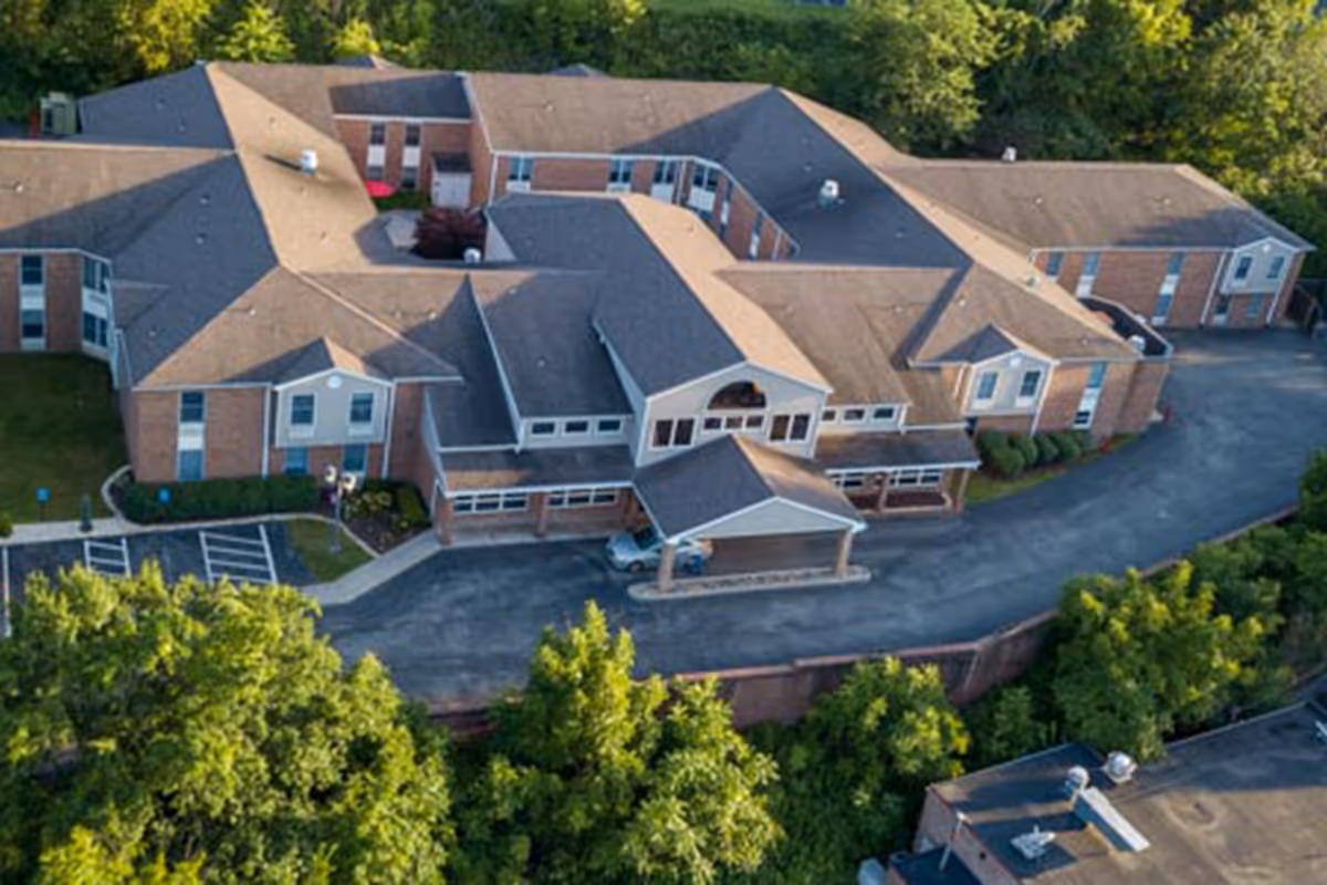 Aerial view of a large brick building with multiple wings and a covered entrance, surrounded by trees and greenery, with a parking area in front.