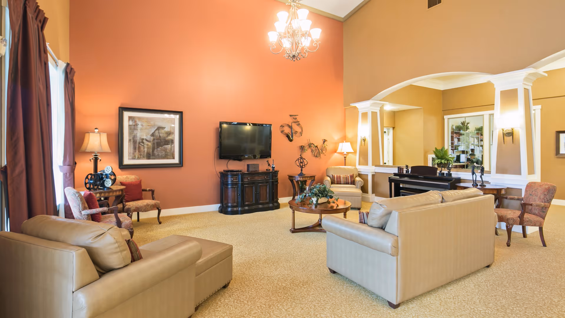 A spacious living room with beige sofas, armchairs, a round wooden coffee table with a plant centerpiece, a flat-screen TV mounted on an orange accent wall, and decorative wall art. The room features a chandelier, large windows with dark curtains, and an open archway leading to another area with a piano and additional seating.