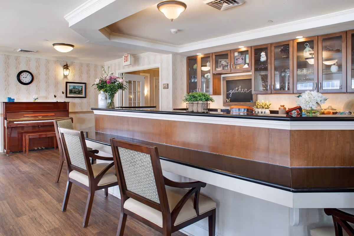 Interior view of a senior living facility common area with a wooden counter and three chairs in the foreground. Behind the counter are glass-front cabinets displaying decorative items and a chalkboard with the word 'gather'. To the left, there is a piano against the wall, a clock, a framed picture, and a vase with flowers on the counter. The room has wood flooring and soft lighting from ceiling fixtures.