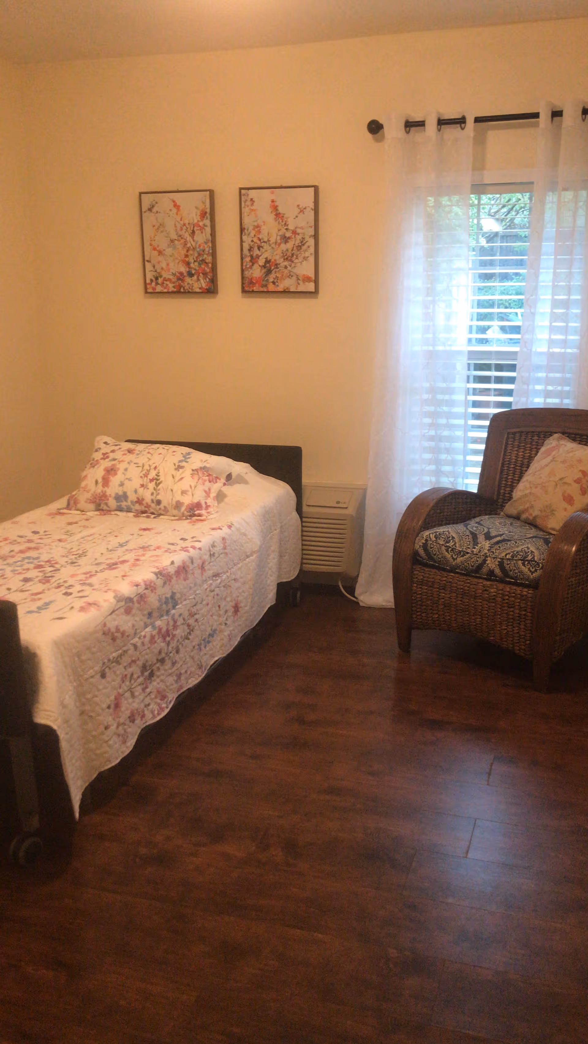 A cozy bedroom with a single bed covered in a floral quilt and matching pillow. Next to the bed is a wicker armchair with a patterned cushion and a floral pillow. Two framed floral artworks hang on the wall above the bed. A window with white sheer curtains allows natural light into the room, and the floor is wooden.