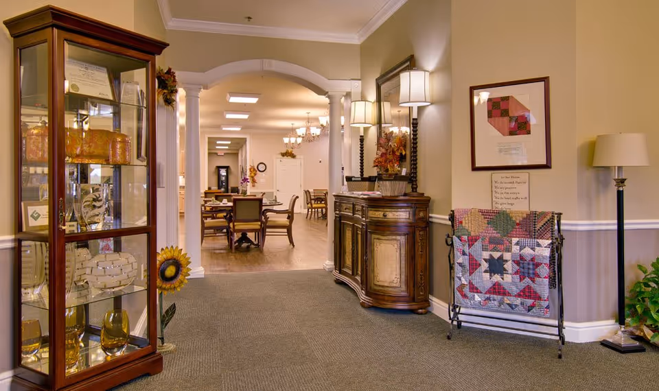 Interior view of a senior living facility hallway with a glass display cabinet on the left containing decorative items, a wooden sideboard with lamps and a mirror on the right, and a quilt rack with a colorful quilt. In the background, there are dining tables and chairs under ceiling lights and chandeliers.