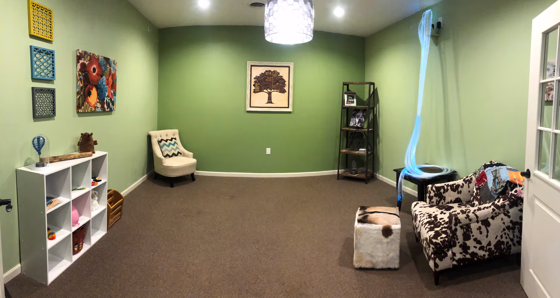 A small room with light green walls and brown carpet. The room contains a white cubby shelf with various items, a beige armchair with a chevron pillow, a framed tree artwork on the wall, a black metal shelf with decorative items, a cow-print armchair with a colorful throw, a small ottoman with a cowhide pattern, and a hanging light fixture with a white shade.