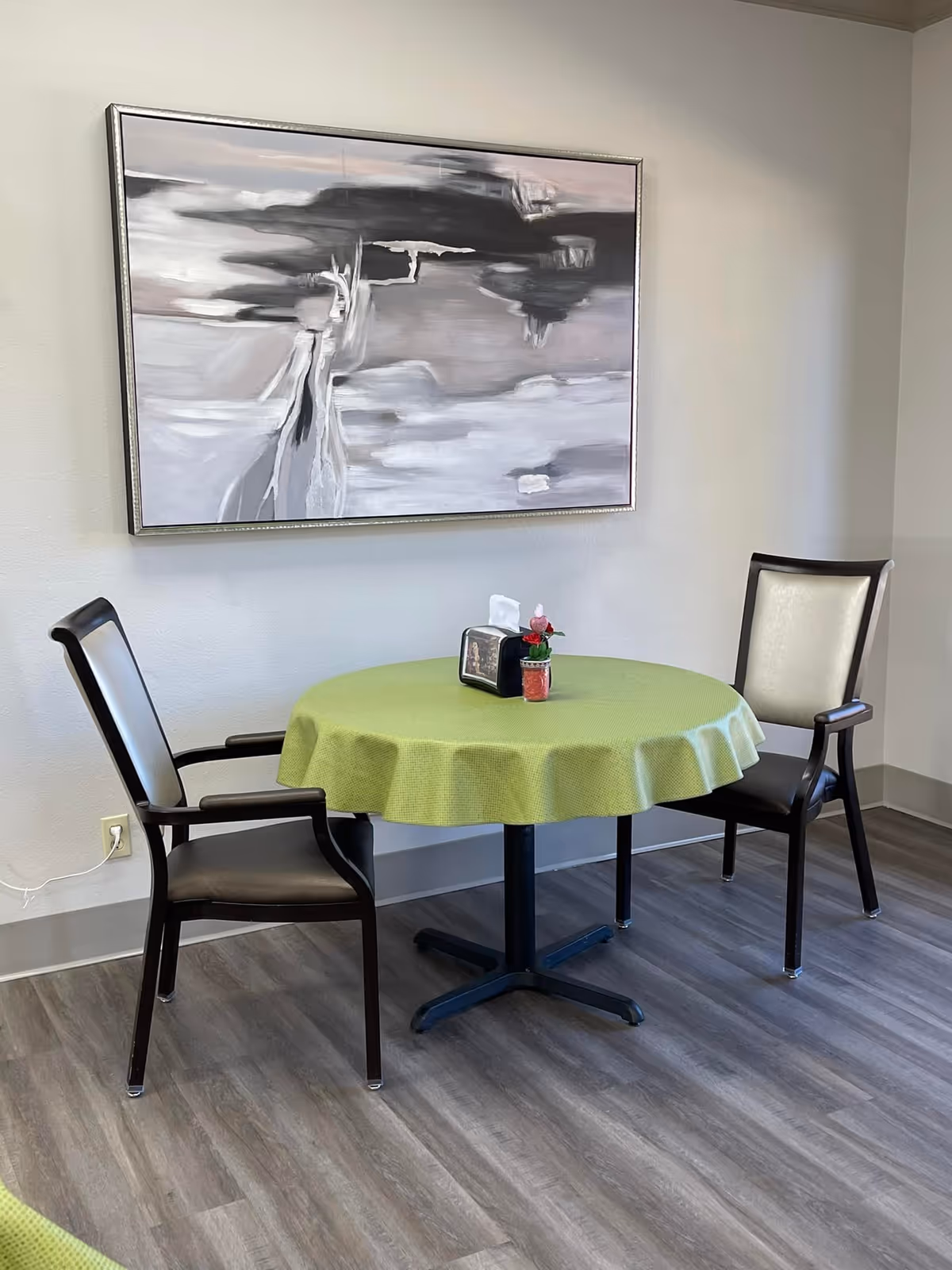 A small round table covered with a green tablecloth is set against a light-colored wall. Two chairs with dark wooden frames and light-colored cushions are positioned on either side of the table. On the table, there is a napkin holder and a small decorative item with a red flower. Above the table hangs a large abstract painting in shades of gray, black, and white. The floor is covered with wood-patterned vinyl or laminate flooring.