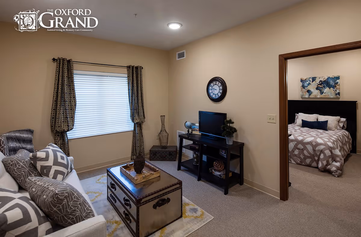 A cozy living room with a light gray sofa adorned with patterned pillows, a decorative trunk-style coffee table on a patterned rug, and a window with geometric patterned curtains. A black TV stand holds a small TV, a globe, and a potted plant. A round wall clock hangs above the TV stand. Through an open doorway, a bedroom is visible with a bed featuring a patterned comforter, pillows, a dark wooden headboard, and a world map artwork on the wall.